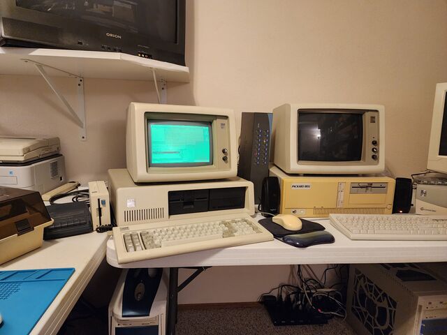 A desk holding an IBM 5150 with a lit green IBM 5151 monitor on top of it showing the os8088 desktop, and beside it a Packard Bell case with an IBM 5153 color monitor on top whose screen is completely dark. A cassette recorder, a printer and other machines fill the rest of the bench.