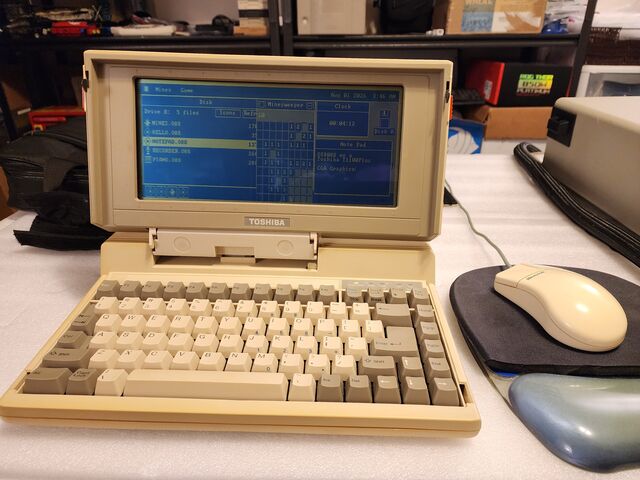 An open Toshiba T1100 Plus laptop on a white bench with a beige Microsoft mouse beside it. Its wide blue LCD shows the os8088 desktop with a Disk window, a Minesweeper board, a Clock and a Note Pad.